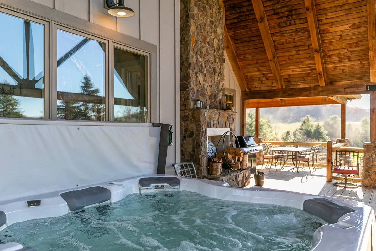 A spacious exterior view featuring a hot tub, a stone fireplace, and an outdoor dining area, all set on a wooden deck surrounded by lush greenery. The scene suggests ample room for socializing, relaxing, and enjoying meals outdoors.