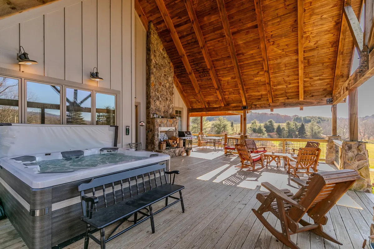 This image features a covered deck area with a hot tub, outdoor seating, and a stone fireplace framed by sturdy wooden beams. The space is open to the natural surroundings, providing panoramic views—a perfect spot for relaxing, socializing, and enjoying the mountain scenery.