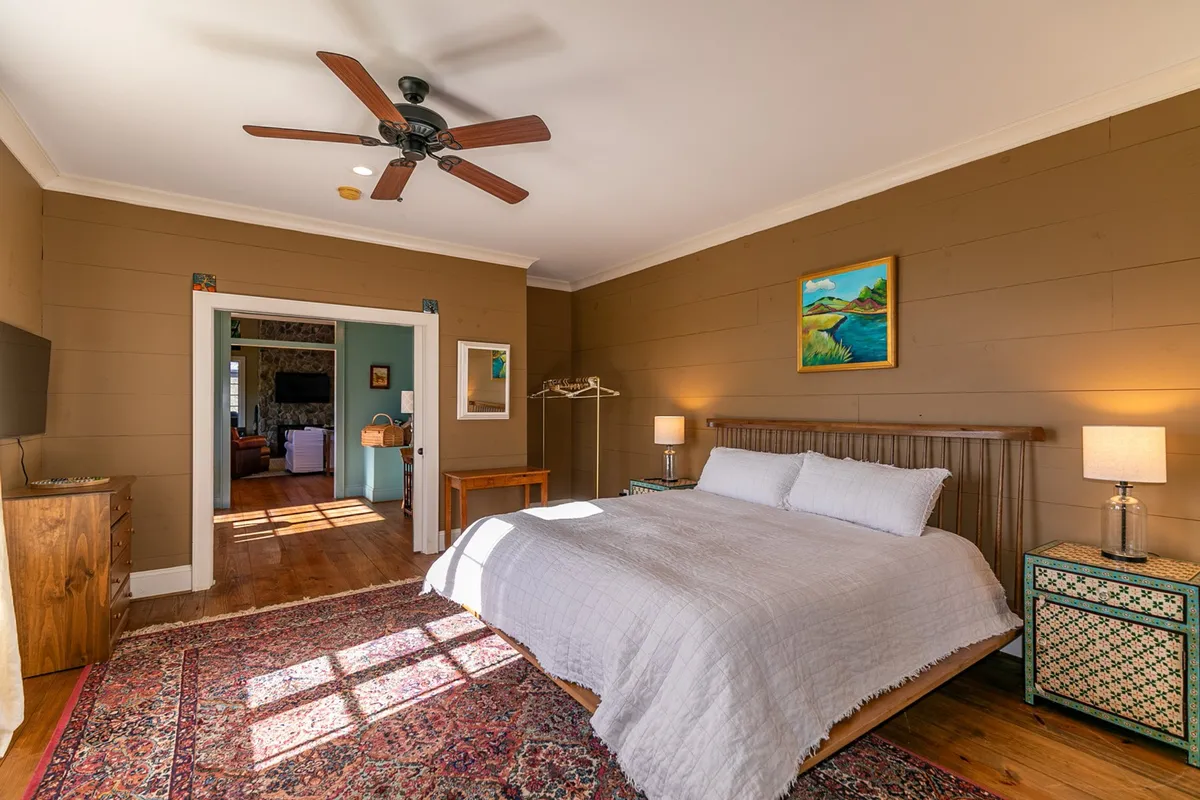 A restful bedroom featuring a plush bed, cozy rug, bedside tables, and soothing artwork. The window lets in soft morning light, creating a warm, welcoming space for relaxation.