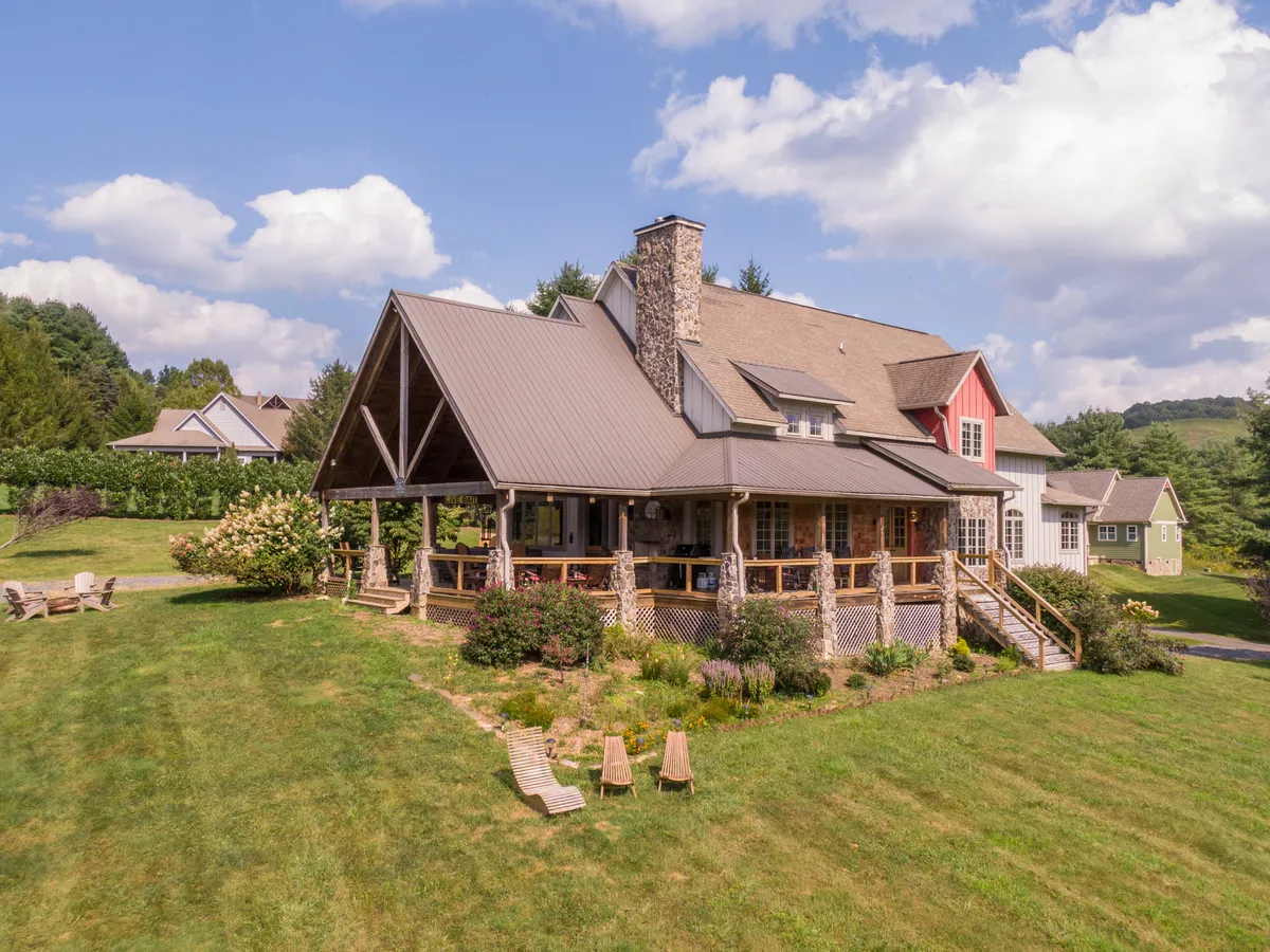 A welcoming view of the large covered porch framed by lush greenery. The scene features a prominent stone chimney, wooden deck chairs, and the tranquil ambiance of a home nestled in nature. The setting suggests a seamless blend between upscale comfort and the outdoors, ideal for relaxing, social gatherings, or simply soaking in the surrounding beauty.