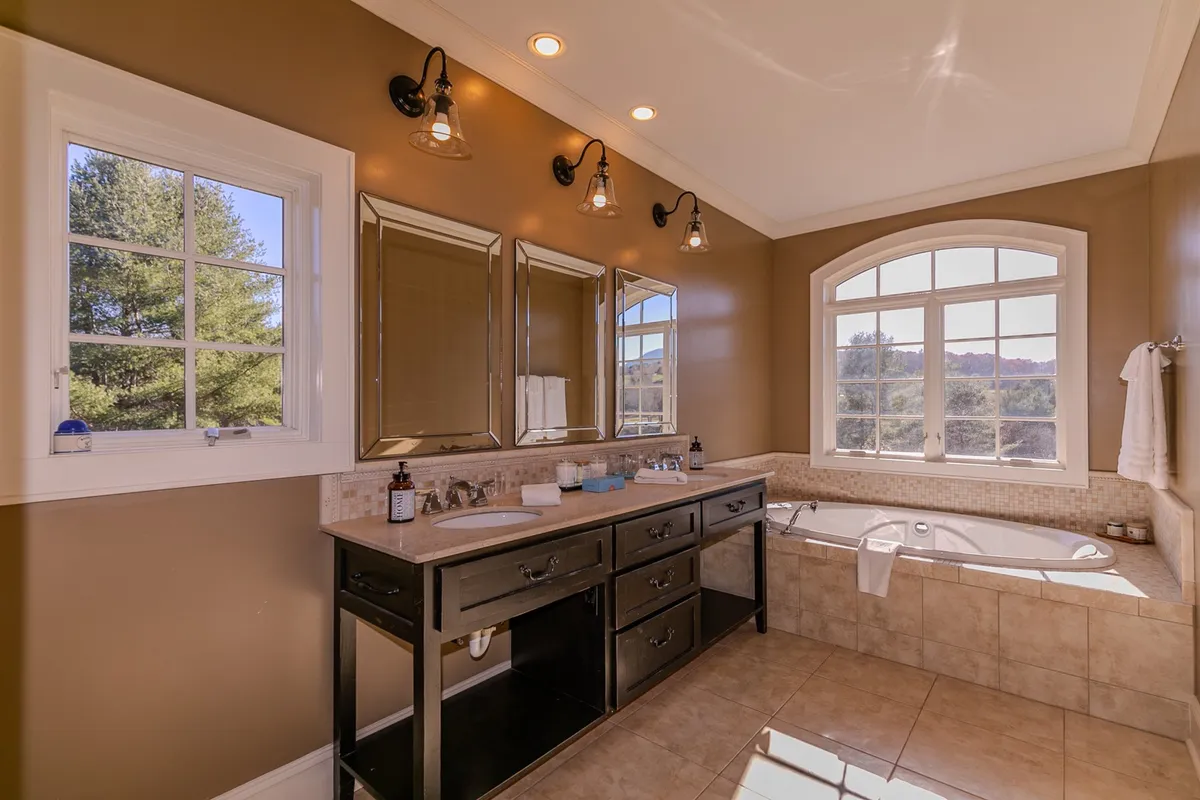 A spacious and luxurious bathroom with a freestanding bathtub, double sink vanity, large mirrors, and arched window allowing in plenty of natural light. The space exudes comfort and relaxation.