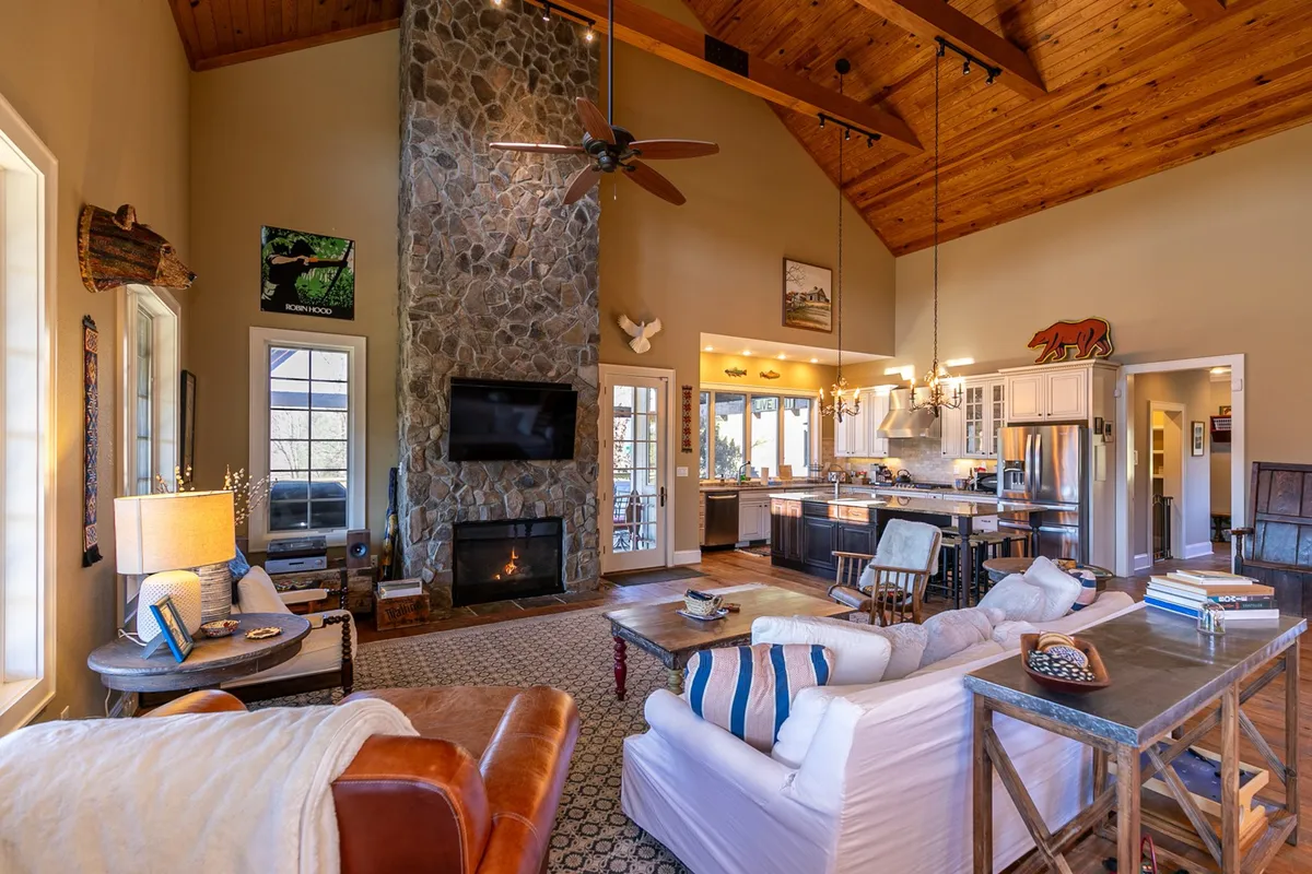 A spacious, sunlit living area with a large stone fireplace reaching up to a high wooden ceiling. Comfortable seating is arranged around the hearth, and a kitchen island is visible nearby, creating an open flow between gathering spaces. Large windows allow ample natural light and connect the space to the outdoors.