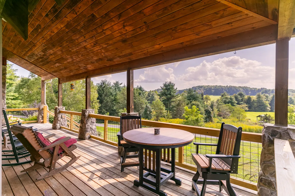 A spacious covered wooden deck with a round table and rocking chairs, offering a scenic view of the mountains and green landscape—seamlessly blending indoor comforts with outdoor beauty.
