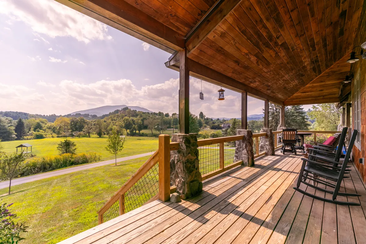 A tranquil exterior shot featuring wooden rocking chairs on a spacious porch, overlooking lush green fields and mountains in the distance. The scene exudes peace and invites guests to relax and soak in the natural surroundings.