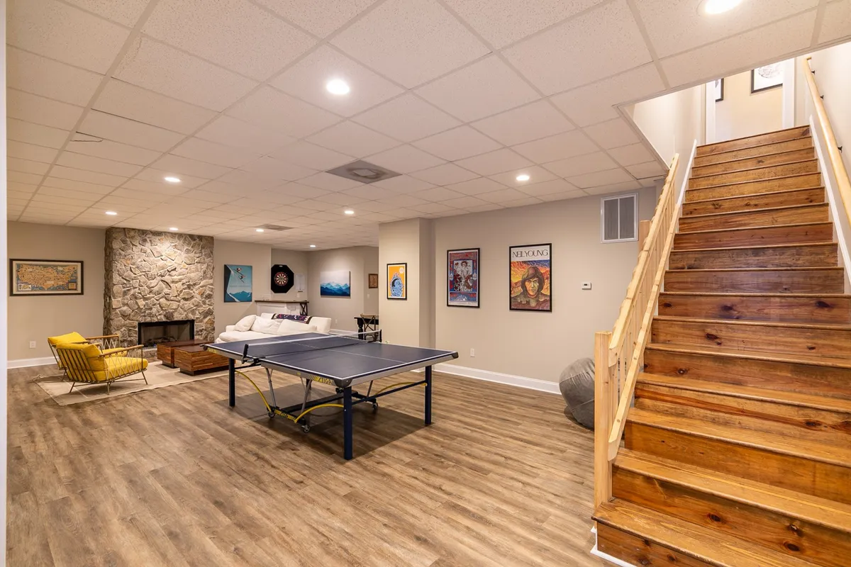 A cozy, open-concept lounge featuring a ping-pong table, a stone fireplace, a yellow armchair, and colorful artwork on the walls. The room is designed for games, relaxation, and movie nights, with a playful yet welcoming vibe.