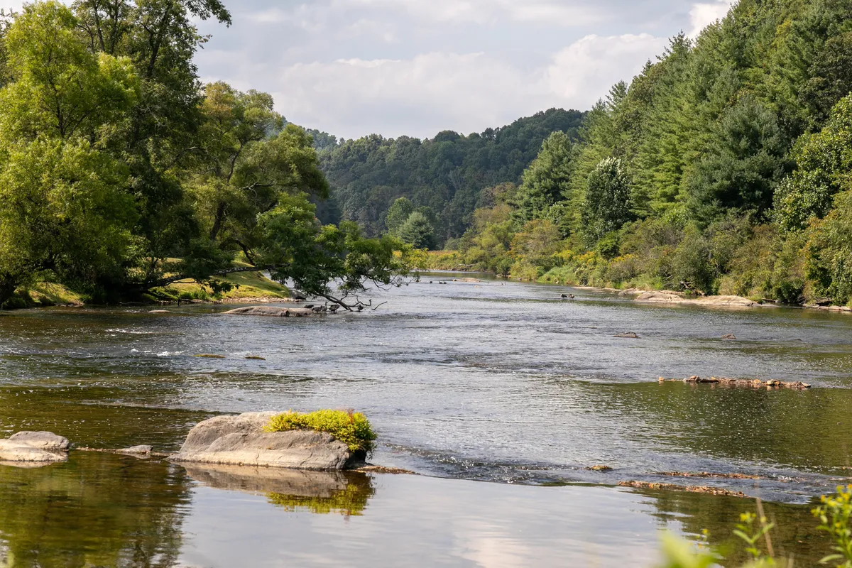 A serene view of the river, framed by lush trees and rock formations along the bank. The tranquil water reflects the sky above, inviting thoughts of kayaking, canoeing, or simply enjoying a peaceful picnic by the riverside.