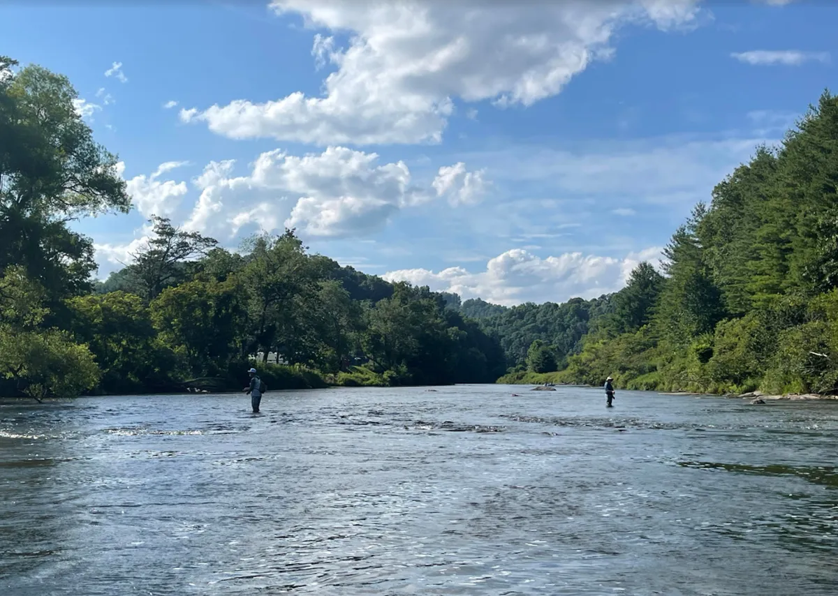 A tranquil river view with fishermen along the banks, trees lining the water, and a gently clouded sky. The peaceful vibe hints at nearby outdoor adventures and group exploration possibilities.