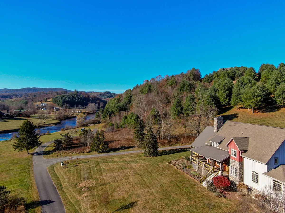 The image showcases a stunning luxury home surrounded by tall trees, with the tranquil New River visible nearby and majestic mountains in the background. The porch offers sweeping sunset views and there's a sense of peaceful seclusion ideal for relaxing or exploring nature.