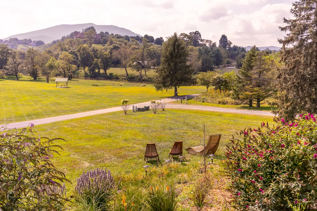 Three wooden chairs rest on a lush green lawn, looking out over flowering plants, a pathway, and distant mountains. The expansive view emphasizes the property's scenic mountain setting, offering a sense of tranquility and adventure. The space is perfect for morning coffee, evening sunsets, or stargazing under open skies.