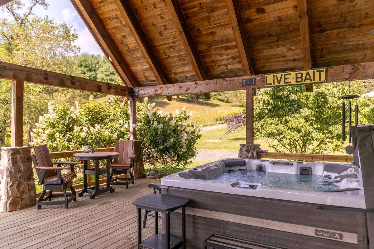 An inviting outdoor space featuring a hot tub on a wooden deck surrounded by greenery, with a dining table nearby and string lights overhead. The area is set up for socializing, relaxing, and enjoying meals al fresco.