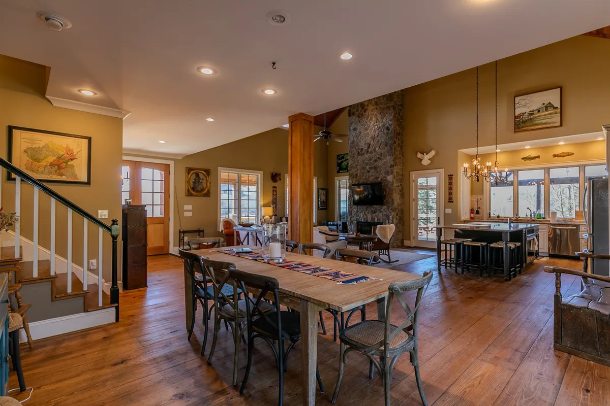 A cozy dining area featuring a sturdy wooden dining table, a stone fireplace adding ambiance, large windows that frame the outdoors, and a staircase in the background. The space is set up for both intimate meals and larger gatherings, with a warm, welcoming mood.